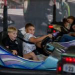 Na zdjęciu widać dwóch chłopców w kolorowych samochodzikach na wesołym miasteczku. Samochodziki są niebieskie i fioletowe, tło jest rozmazane. Chłopcy wyglądają na zadowolonych i skoncentrowanych.