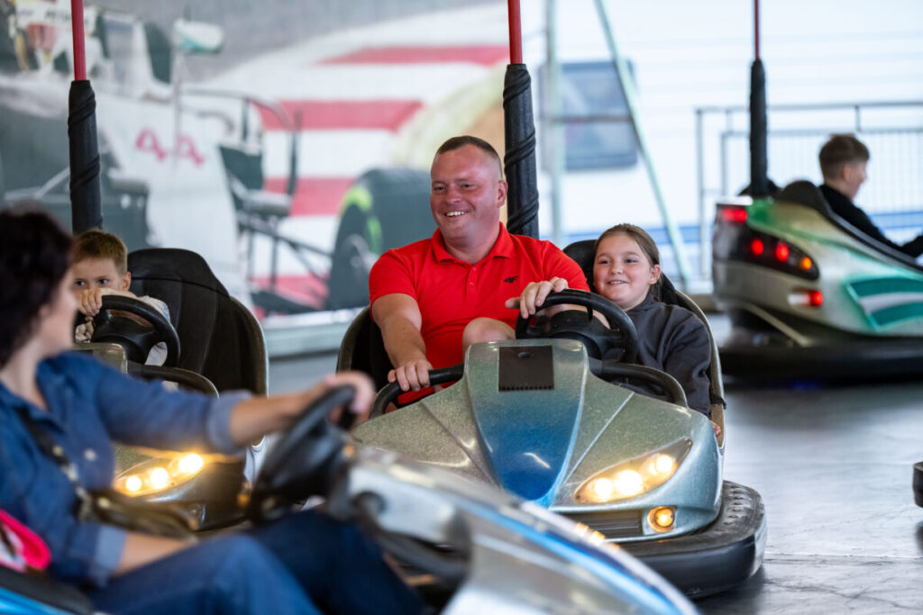 Na zdjęciu widać osoby bawiące się na autodromie. W centrum mężczyzna w czerwonej koszulce z dzieckiem. W tle widać wesołe kolory i inne zderzające się samochodziki.