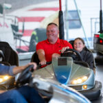 Na zdjęciu widać osoby bawiące się na autodromie. W centrum mężczyzna w czerwonej koszulce z dzieckiem. W tle widać wesołe kolory i inne zderzające się samochodziki.