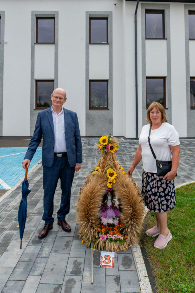 Starszy mężczyzna i kobieta stoją przy ozdobie z kłosów i słoneczników. Tło to biały budynek. Ozdoba jest kolorowa i ma tabliczkę z numerem trzy.