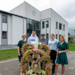 Grupa ludzi stoi przed nowoczesnym białym budynkiem z zegarem. Przed nimi piękny wieniec z kwiatów i kłosów. W tle powiewa polska flaga.