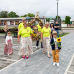 Grupa ludzi w kolorowych strojach idzie chodnikiem. Niosą ozdobne wieńce, widoczne słoneczniki. W tle budynki i pochmurne niebo.