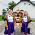 Dwóch kobiet w fioletowych spódnicach i białych bluzkach stoi przed dekoracją z kwiatów i zbóż. Tło to białe budynki i zielone drzewa. Obraz ma jasne, naturalne oświetlenie.