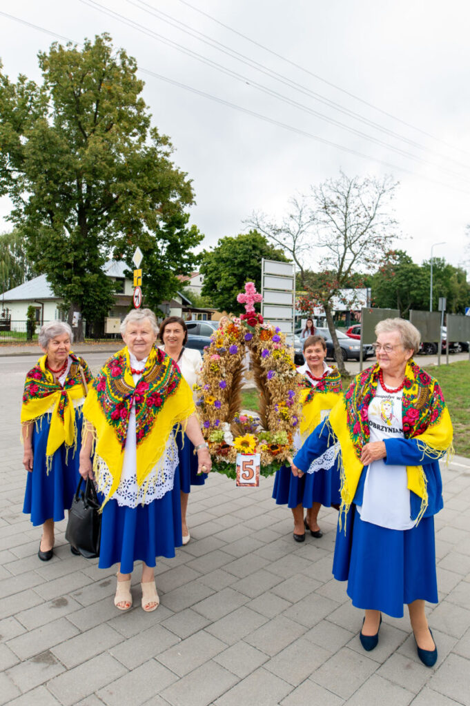 Grupa kobiet w kolorowych strojach ludowych stoi na tle drzew. Mają żółte chusty i niebieskie spódnice. Trzymają ozdobną koronę z kwiatów.