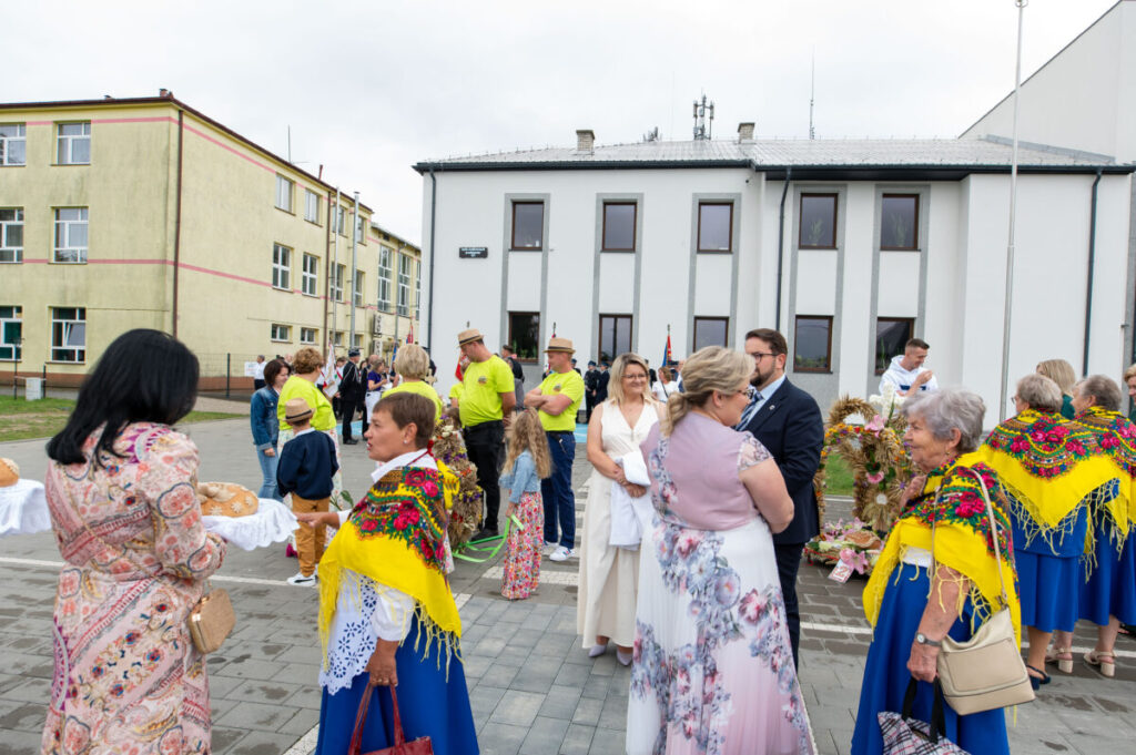 Grupa ludzi w tradycyjnych strojach ludowych w żywych kolorach przed białym budynkiem. Tło to szkoła i szare niebo. Uczestnicy trzymają chleb na tacy.