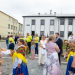 Grupa ludzi w tradycyjnych strojach ludowych w żywych kolorach przed białym budynkiem. Tło to szkoła i szare niebo. Uczestnicy trzymają chleb na tacy.