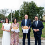 Cztery osoby na tle zieleni, trzymają dekoracyjne chleby na białych serwetach. Niebo pochmurne. Eleganckie ubrania.