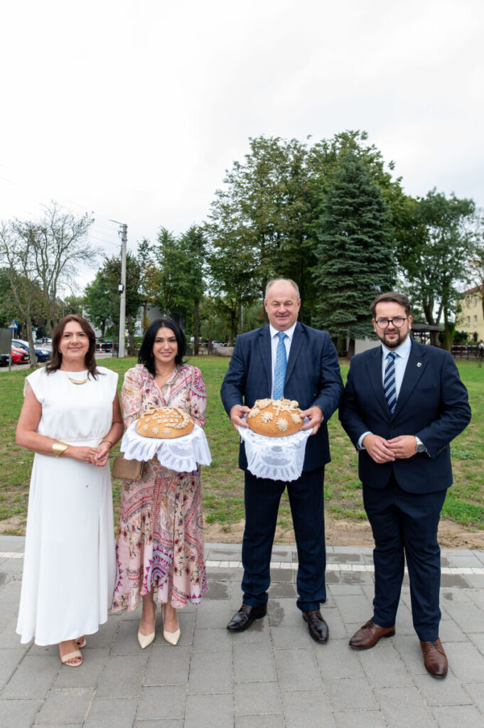 Cztery osoby na tle zieleni, trzymają dekoracyjne chleby na białych serwetach. Niebo pochmurne. Eleganckie ubrania.