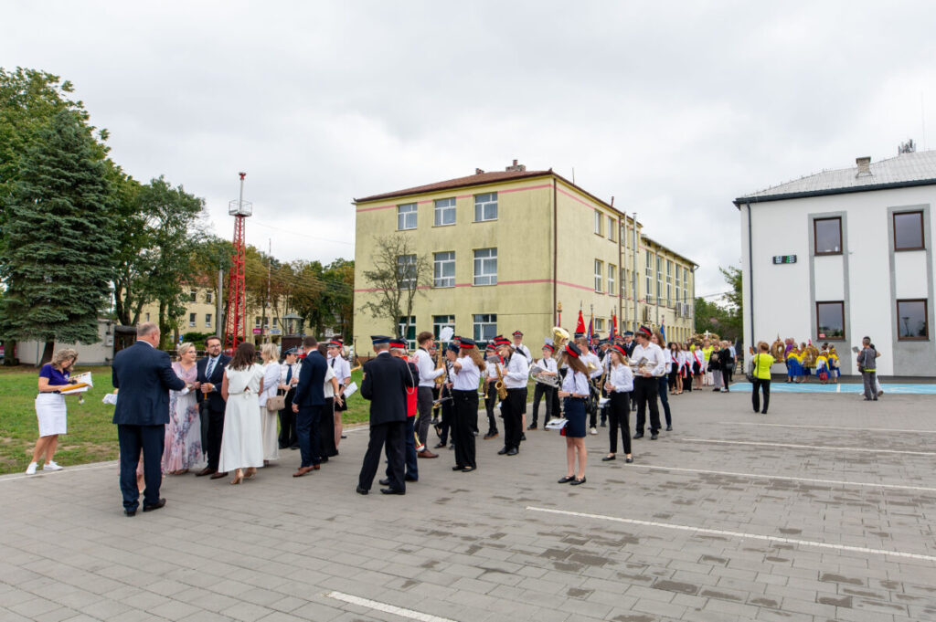 Na zdjęciu widać grupę ludzi w eleganckich strojach na placu, przed budynkami. Dominuje biel, czerń i czerwone akcenty. W tle drzewa i szare niebo.