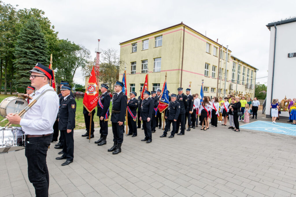 Na zdjęciu jest parada strażaków w mundurach, niosących czerwone sztandary. W tle widoczne jest szkolne, żółte budynek. Obok maszerują ludzie w eleganckich strojach.