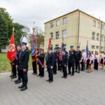 Uroczysty pochód strażaków w mundurach z czerwonymi sztandarami na tle żółtego budynku. W tle drzewa i trawnik. Pogoda pochmurna.