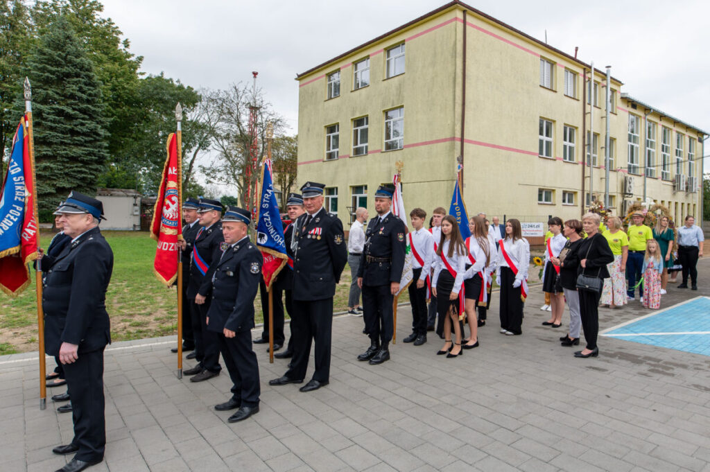 Grupa osób w uniformach i białych koszulach stoi przed budynkiem z flagami. Tło to żółty budynek i drzewa. Uroczysty charakter.