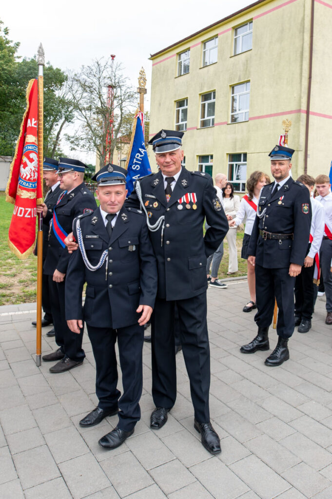 Grupa strażaków w czarnych mundurach stoi przed budynkiem. Tło: kremowy budynek, drzewa. Widoczne są kolorowe sztandary.