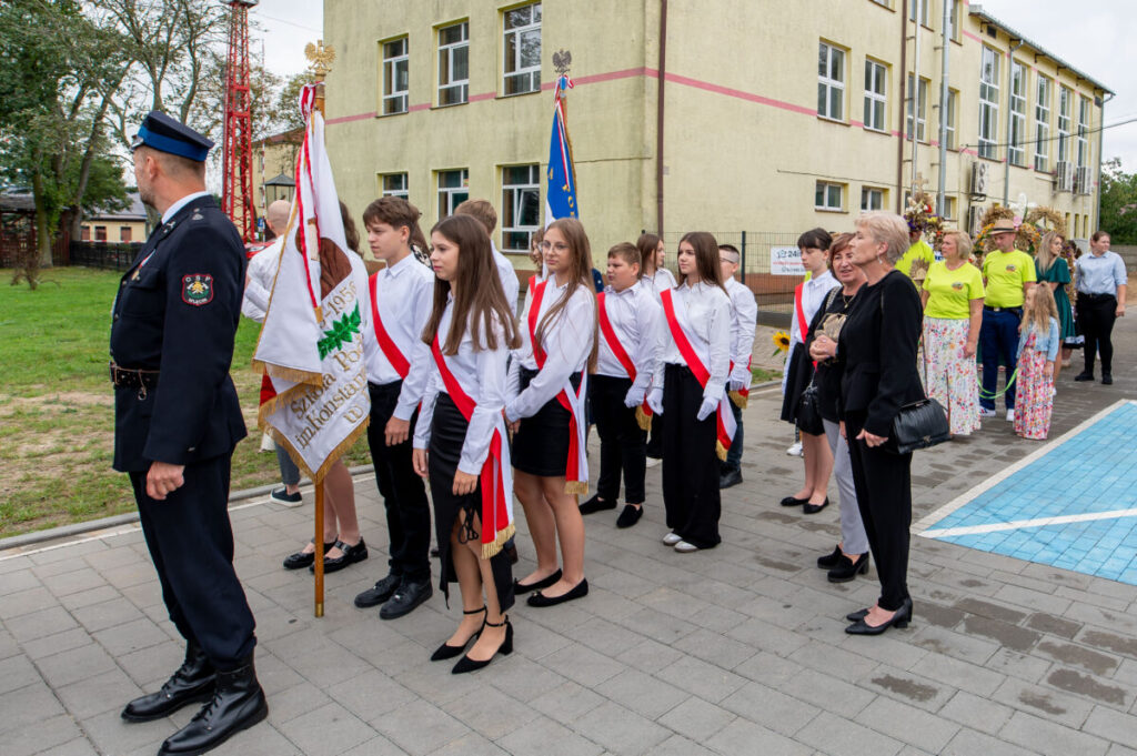 Grupa dzieci w białych koszulach i czerwonych szarfach stoi przed budynkiem szkoły. W tle są dorośli i inne osoby w kolorowych strojach. Dzieci trzymają sztandary szkolne, tworząc oficjalny nastrój.