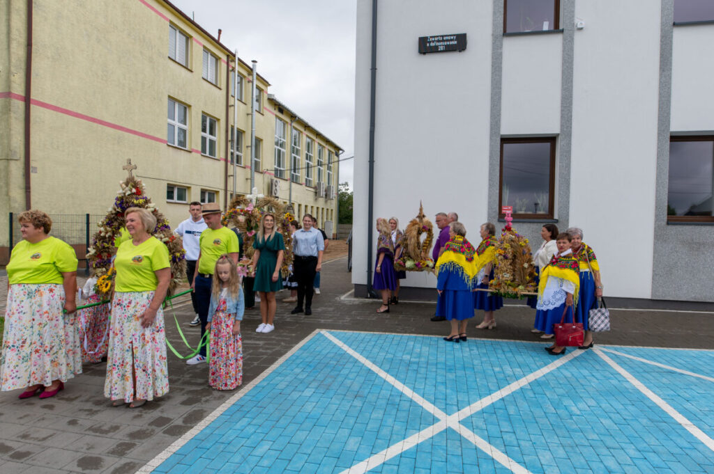 Grupa osób w kolorowych strojach na tle budynku. Dekoracje z kwiatów i zboża. Niebieskie płytki na pierwszym planie.