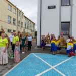 Grupa osób w kolorowych strojach na tle budynku. Dekoracje z kwiatów i zboża. Niebieskie płytki na pierwszym planie.