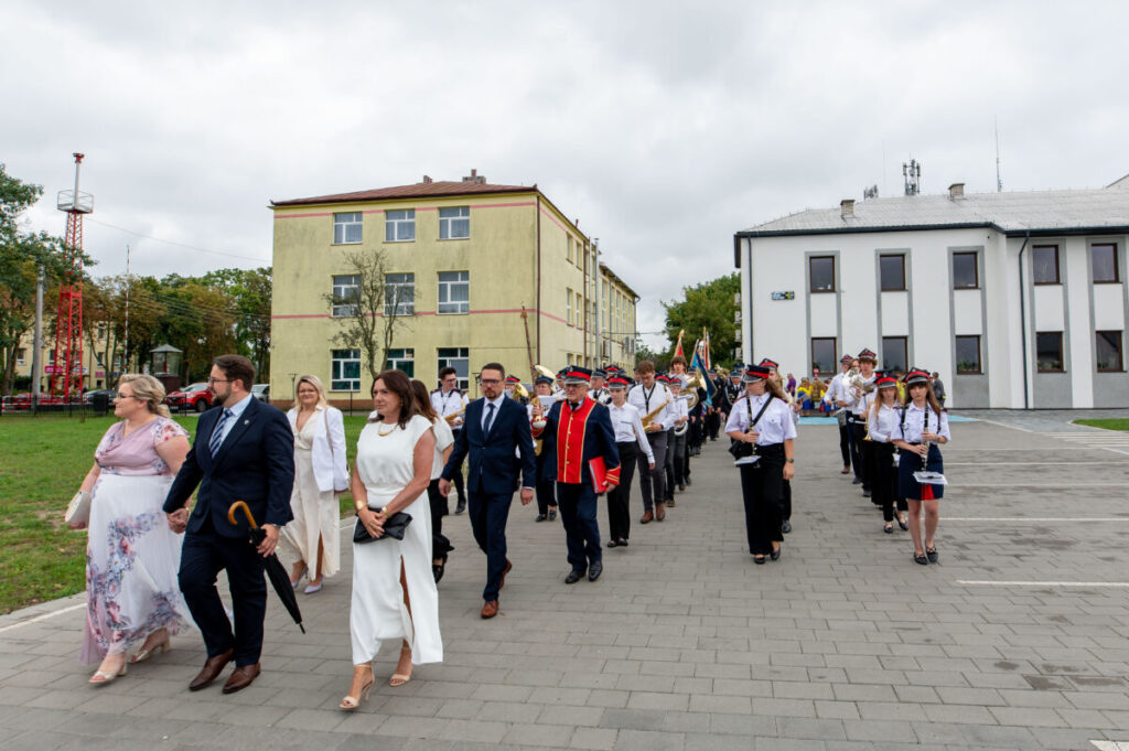 Grupa elegancko ubranych ludzi idzie ulicą w towarzystwie orkiestry dętej. W tle jasnożółty budynek z kilkoma oknami. Dzień jest pochmurny, a osoby wyglądają na zadowolone.