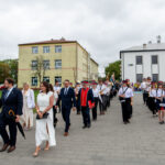 Grupa elegancko ubranych ludzi idzie ulicą w towarzystwie orkiestry dętej. W tle jasnożółty budynek z kilkoma oknami. Dzień jest pochmurny, a osoby wyglądają na zadowolone.