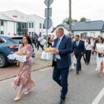 Ludzie w eleganckich strojach idą ulicą, niosąc chleb na białych serwetach. W tle orkiestra i budynki w bieli. Obok stoi niebieski samochód.