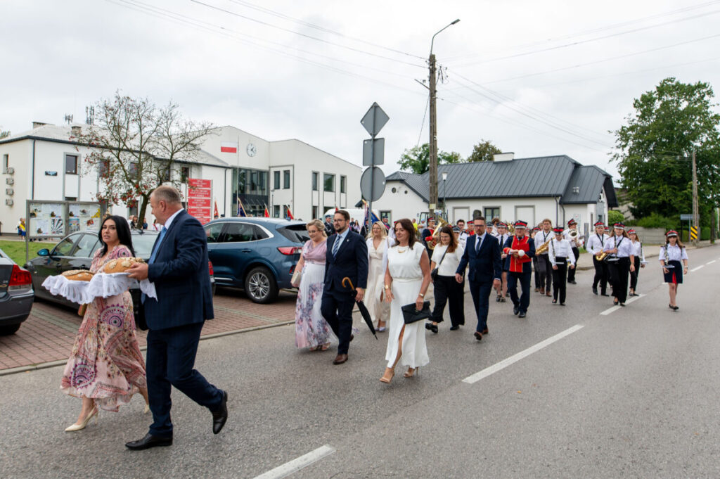 Parada ludzi w eleganckich strojach idących ulicą przy budynku. Mężczyzna niesie chleb na białej serwetce. Tło z samochodami, drzewami i budynkami.