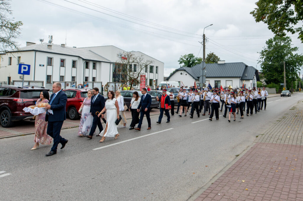 Grupa ludzi w eleganckich strojach i orkiestra maszerują ulicą. Budynki w tle są białe. Pogoda jest pochmurna.