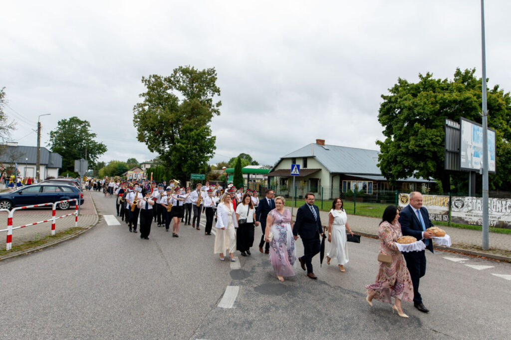 Grupa ludzi maszeruje ulicą w jasnych strojach. W tle widoczne są drzewa i budynki. Niebo jest pochmurne.