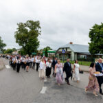 Grupa ludzi maszeruje ulicą w jasnych strojach. W tle widoczne są drzewa i budynki. Niebo jest pochmurne.