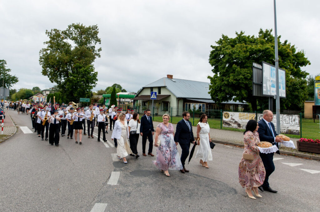 Ludzie idą w procesji po ulicy, w towarzystwie orkiestry dętej. Ubrani elegancko, jasne i ciemne kolory. W tle budynek, drzewa.