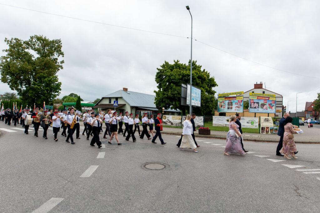 Pochód orkiestry ulicznej w czarnych spódnicach i białych koszulach na szarej drodze. W tle zielone drzewa i budynek. Obok przechodzą elegancko ubrani ludzie.