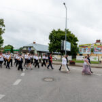 Pochód orkiestry ulicznej w czarnych spódnicach i białych koszulach na szarej drodze. W tle zielone drzewa i budynek. Obok przechodzą elegancko ubrani ludzie.
