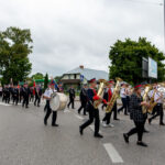 Parada uliczna z orkiestrą dętą w mundurach. Tło: drzewa, budynek i billboard. Niebo pochmurne, droga asfaltowa.