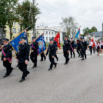 Strażacy w mundurach maszerują z flagami na szarej ulicy. Otoczenie zieleni i trudne niebo. Tło miejskie z drzewami.