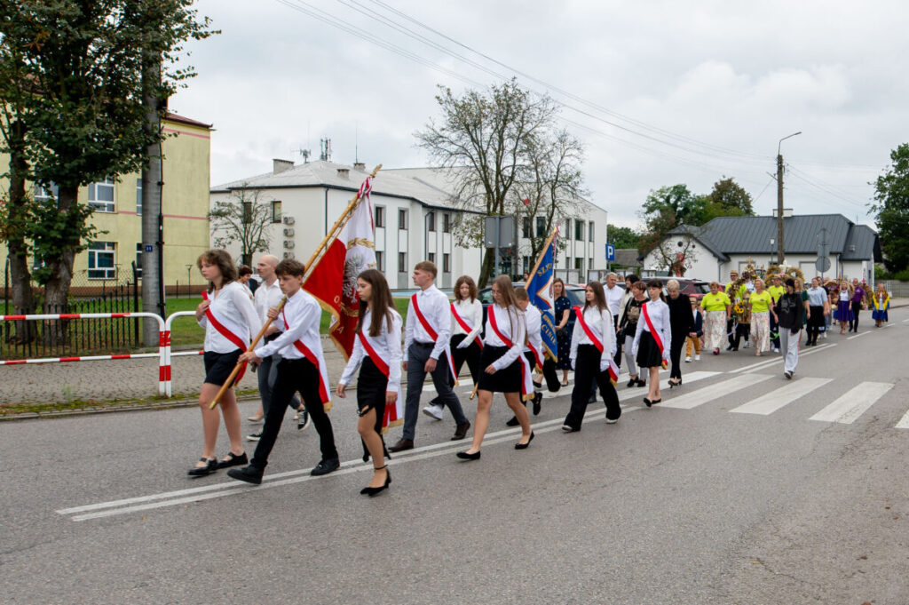 Grupa ludzi w białych koszulach z czerwonymi szarfami maszeruje po ulicy, niosąc flagi. Tło to budynki i drzewa w pochmurny dzień. Kolory dominujące to biel, czerwień i zieleń.