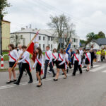 Grupa ludzi w białych koszulach z czerwonymi szarfami maszeruje po ulicy, niosąc flagi. Tło to budynki i drzewa w pochmurny dzień. Kolory dominujące to biel, czerwień i zieleń.