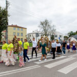 Grupa ludzi w kolorowych strojach idzie wzdłuż ulicy. Tło stanowią budynki i drzewa. Atmosfera sugeruje lokalne święto.