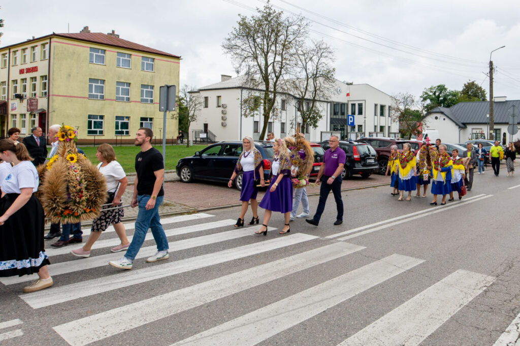 Na zdjęciu widać grupę osób w kolorowych strojach ludowych, przechodzącą przez przejście dla pieszych. Tło stanowią budynki mieszkalne i zaparkowane samochody. Widać też tradycyjne wieńce z kwiatów i zbóż.