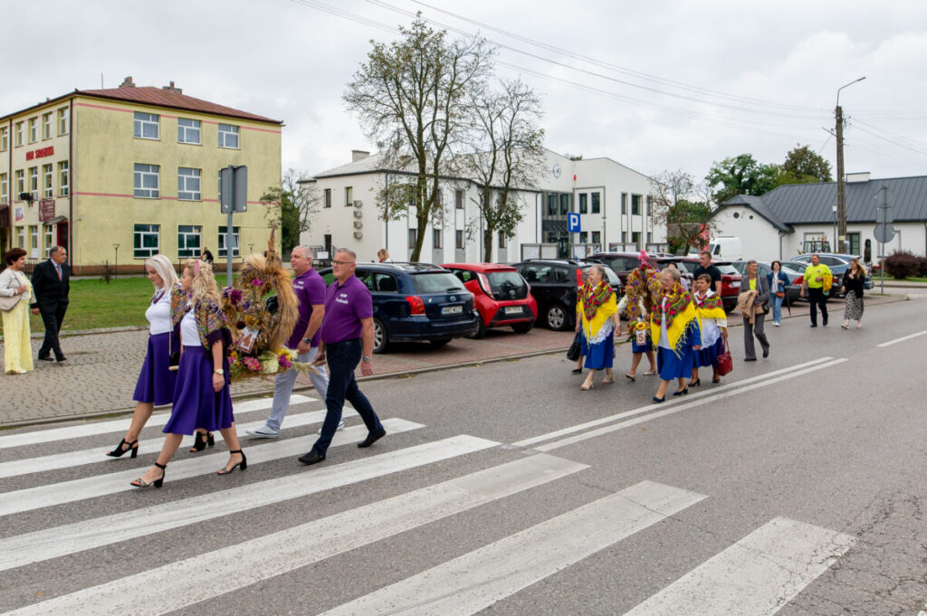 Grupa ludzi w strojach ludowych przechodzi przez przejście. Ubrania w fioletach i kolorowe szale. W tle budynki i zaparkowane auta.