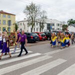 Grupa ludzi w strojach ludowych przechodzi przez przejście. Ubrania w fioletach i kolorowe szale. W tle budynki i zaparkowane auta.