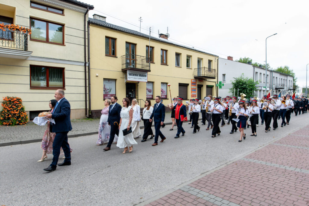 Parada na ulicy: ludzie w eleganckich strojach, orkiestra w białych koszulach i czerwonych mundurach. Tło kamienic.
