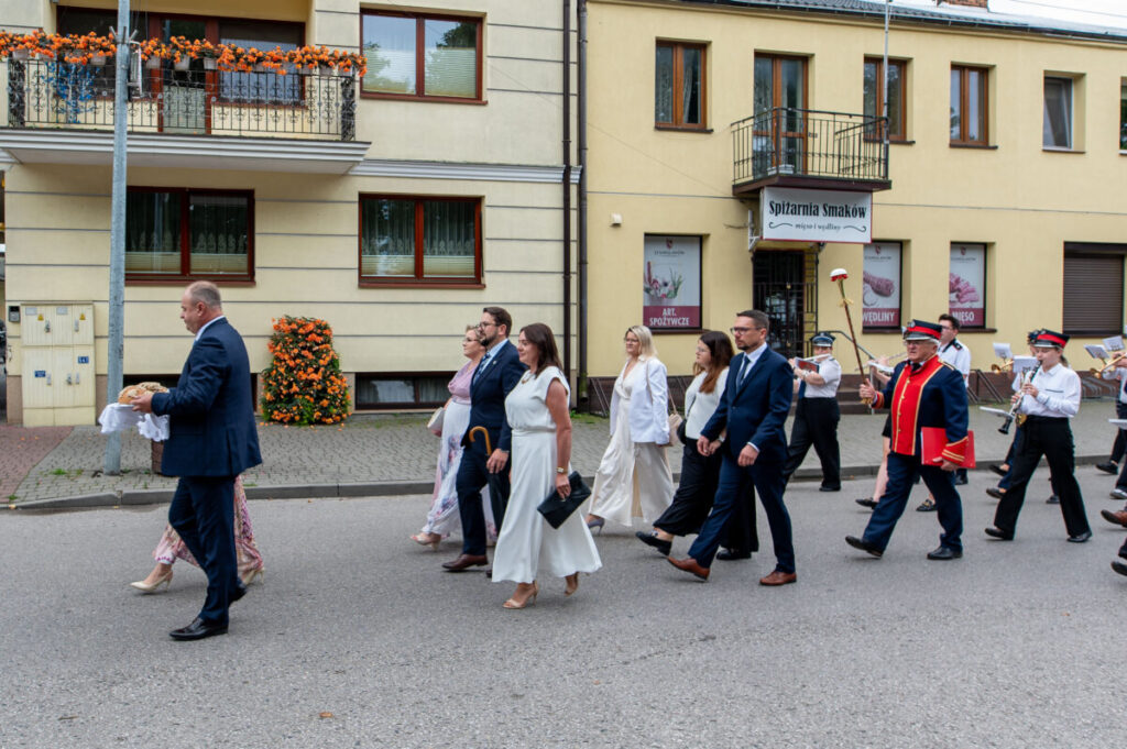 Grupa ludzi w eleganckich strojach idzie ulicą w paradzie. Na czele mężczyzna niesie chleb na białej serwecie. Tło stanowią jasne budynki z oknami ozdobionymi kwiatami.