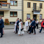 Grupa ludzi w eleganckich strojach idzie ulicą w paradzie. Na czele mężczyzna niesie chleb na białej serwecie. Tło stanowią jasne budynki z oknami ozdobionymi kwiatami.