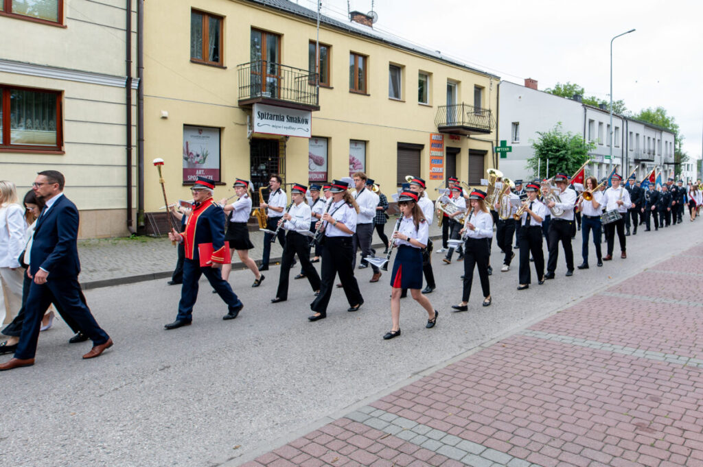 Orkiestra maszeruje ulicą w białych koszulach i granatowych mundurach, grając na instrumentach dętych. Tło stanowi żółty budynek z balkonami. Na chodniku widać czerwono-szarą kostkę brukową.