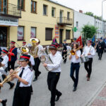 Orkiestra dęta maszeruje ulicą, ubrana w białe koszule i czerwone czapki. W tle żółty budynek z szyldem sklepu. Po prawej widoczni strażacy i przechodnie.