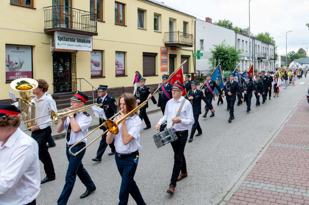 Orkiestra marszowa gra na ulicy obok budynku w kremowym kolorze. Uczestnicy noszą białe koszule i czarne spodnie. W tle powiewają barwne flagi.