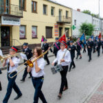 Orkiestra marszowa gra na ulicy obok budynku w kremowym kolorze. Uczestnicy noszą białe koszule i czarne spodnie. W tle powiewają barwne flagi.