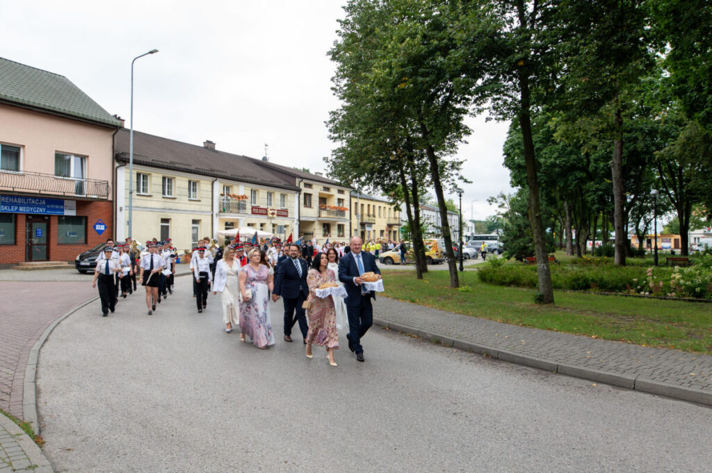 Grupa ludzi w eleganckich strojach idzie ulicą. Towarzyszy im orkiestra w mundurach. W tle widoczne są budynki i drzewa.