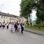 Grupa ludzi w eleganckich strojach idzie ulicą. Towarzyszy im orkiestra w mundurach. W tle widoczne są budynki i drzewa.