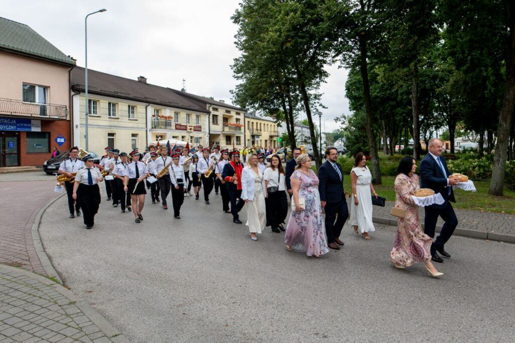 Na zdjęciu widać marsz ludzi w eleganckich strojach i orkiestrę. Uczestnicy niosą chleb na białych serwetach. Tło to budynki i drzewa, a niebo jest pochmurne.