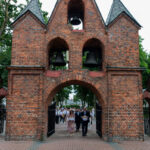 Ceglana brama z trzema dzwonami, przed nią idą ludzie. Drzewa w tle dodają zieleni. Brukowana ścieżka prowadzi przez bramę.