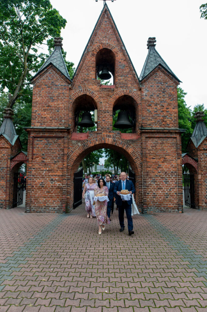 Ceglana brama z dzwonami, pod nią ludzie elegancko ubrani, otoczeni zielenią. Tło to drzewa i jasne niebo. Na brickowym bruku kroczą grupa.